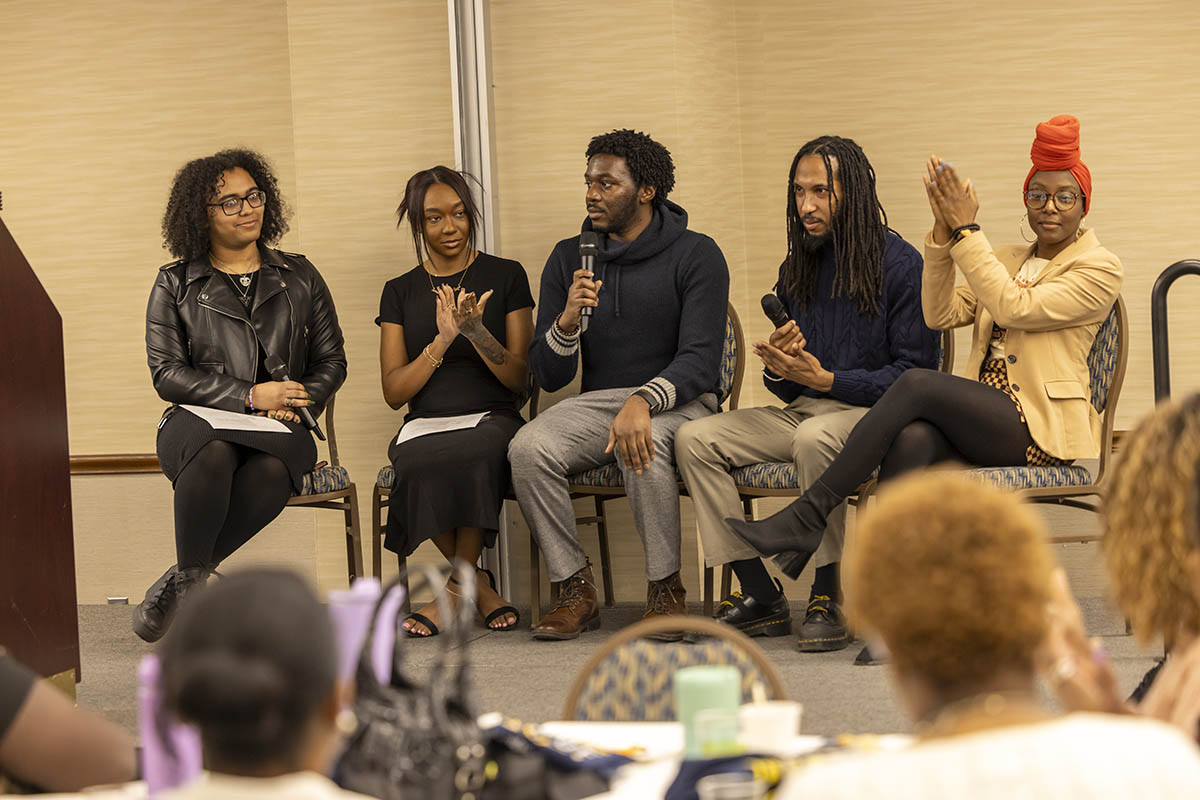 A panel of UM-Flint students seated on stage speaking into a microphne