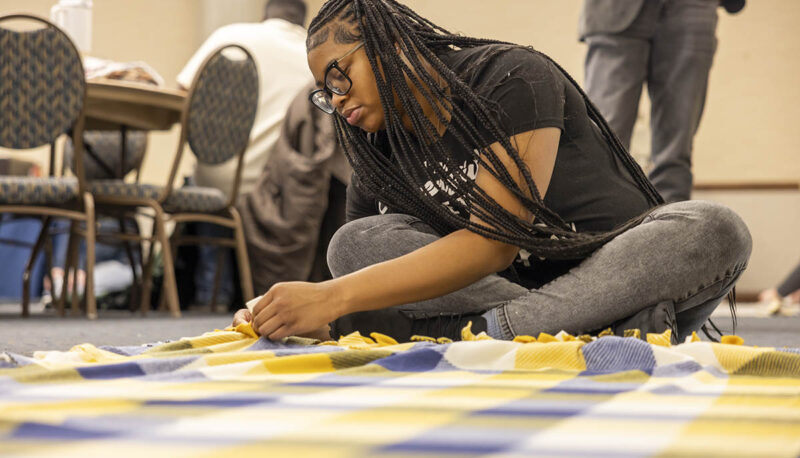 A UM-Flint volunteer seated on the floor tying a blanket.