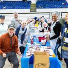 Donna Fry stands with a group of volunteers around a table prepping food in a large gym space, there are packages of food around on the ground.