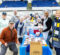 Donna Fry stands with a group of volunteers around a table prepping food in a large gym space, there are packages of food around on the ground.