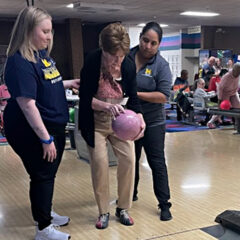 Two female UM OT students assist a stroke patient with bowling. Patient holds a light pink bowling ball.