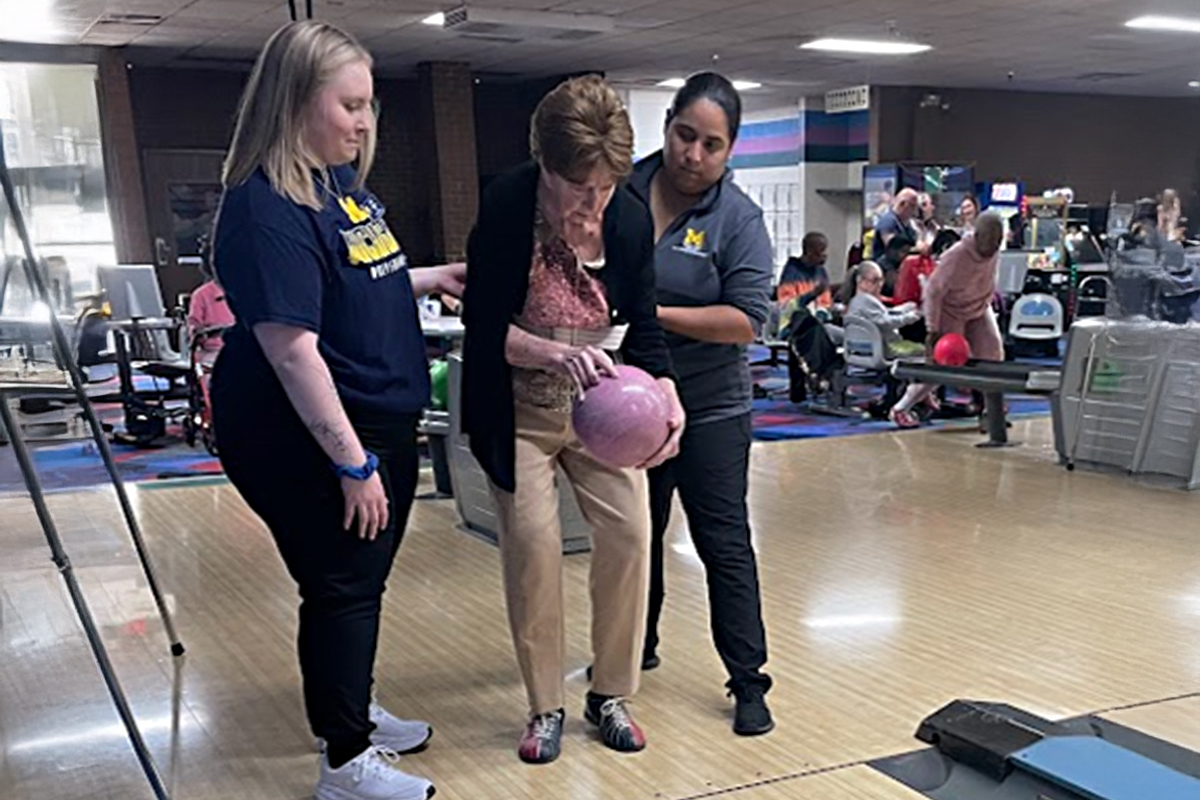 Two female UM OT students assist a stroke patient with bowling. Patient holds a light pink bowling ball.