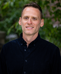 Portrait of a man in a dark shirt with green trees and leaves in the background