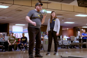 An occupational therapy patient getting ready to throw a bowling ball down the lane at an alley while an OT student stands behind him.