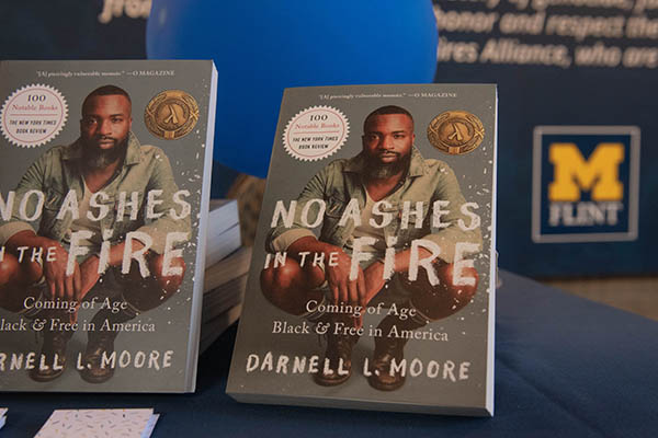 Two copies of "No Ashes in the Fire" on a table with a wall featuring the UM-Flint logo in the background. 