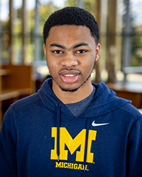 A portrait of a young Black man wearing a University of Michigan sweatshirt smiling at the camera