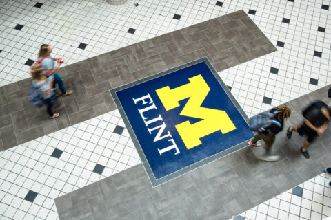 An overhead photo of the main floor of the University Pavilion, showing students walking on the tile floor and the UM-Flint logo in tile.
