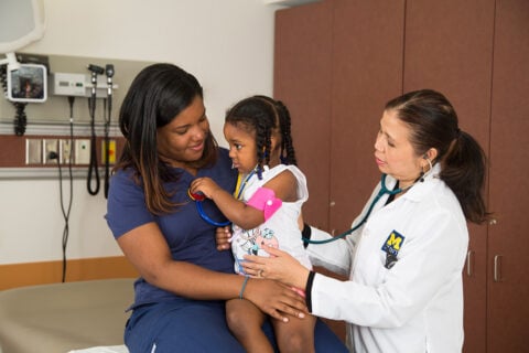 A UM-Flint health care provide checking a young girl sitting on her mom's lap.