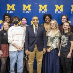 A group of staff and students posing in front of a UM-Flint banner during Lavender Graduation. Chancellor Laurence Alexander is front and center.