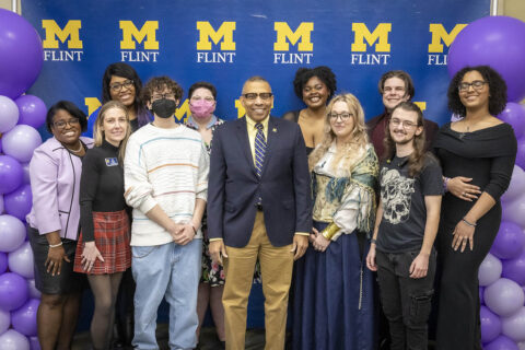 A group of staff and students posing in front of a UM-Flint banner during Lavender Graduation. Chancellor Laurence Alexander is front and center.