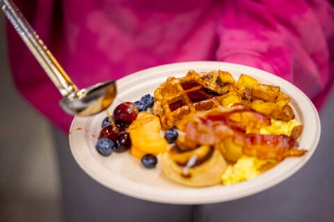 A plate with a waffle, bacon, eggs, and fruit with syrup being ladled over it.