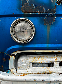 A close-up photograph of a bright blue automobile's headlight with rust deterioration and stains