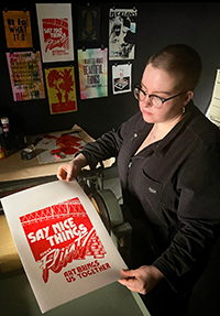 A portrait of Janice McCoy, a young woman with a shaved head holding a letterpress print, with more prints hanging on the wall behind her.