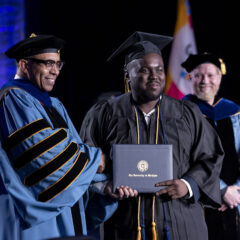 A student receiving his diploma on stage, shaking Chancellor Laurence Alexander's hand. Interim Provost Yener Kandogan looks on in the background.