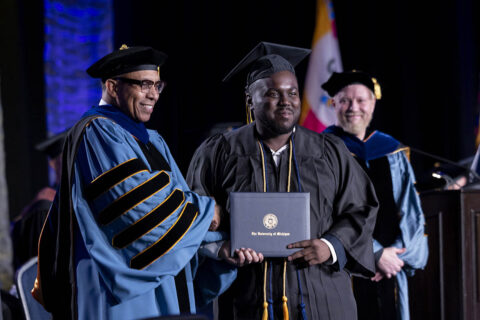 A student receiving his diploma on stage, shaking Chancellor Laurence Alexander's hand. Interim Provost Yener Kandogan looks on in the background.