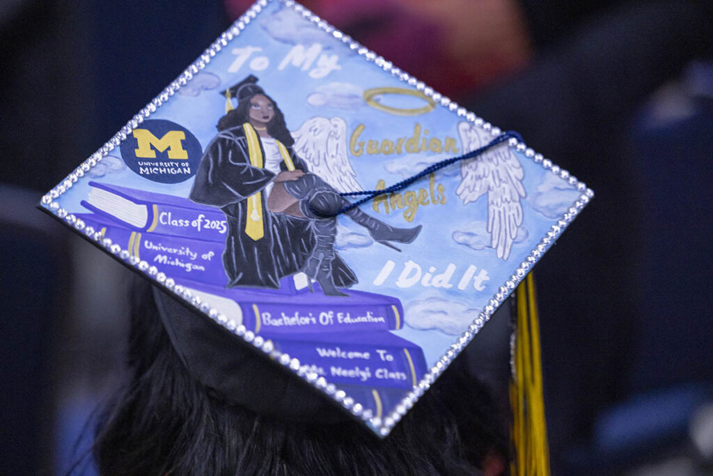 A painted graduation cap. The painted shows an African American woman in a graduation cap sitting on steps of books. Text says: To my Guardian Angels. I Did It.