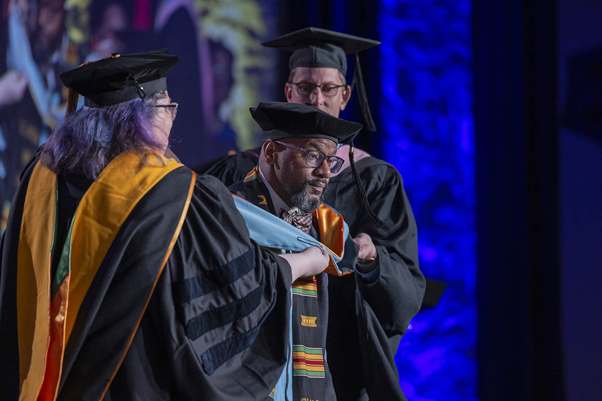 A student being arrayed in his graduate hood on stage.