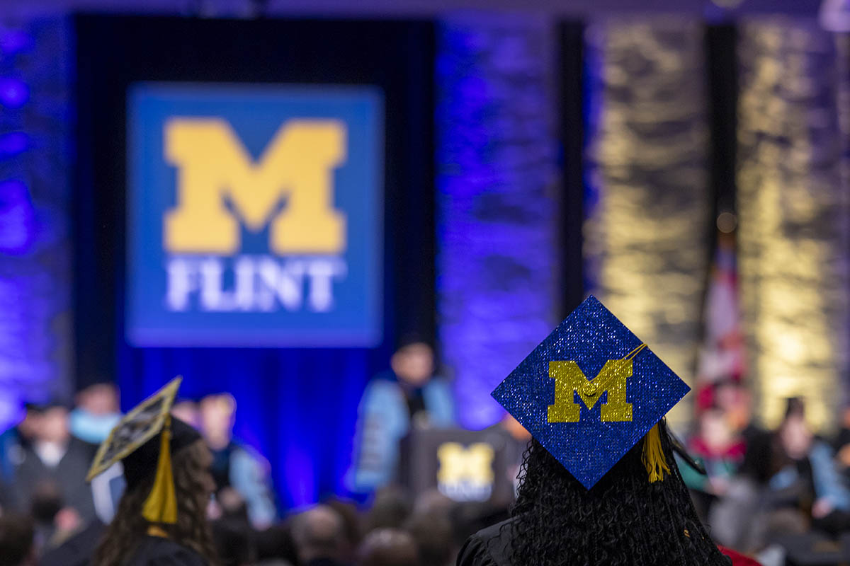 A graduation cap decorated with the Block Michigan "M" with the UM-Flint stamp logo being displayed on stage.