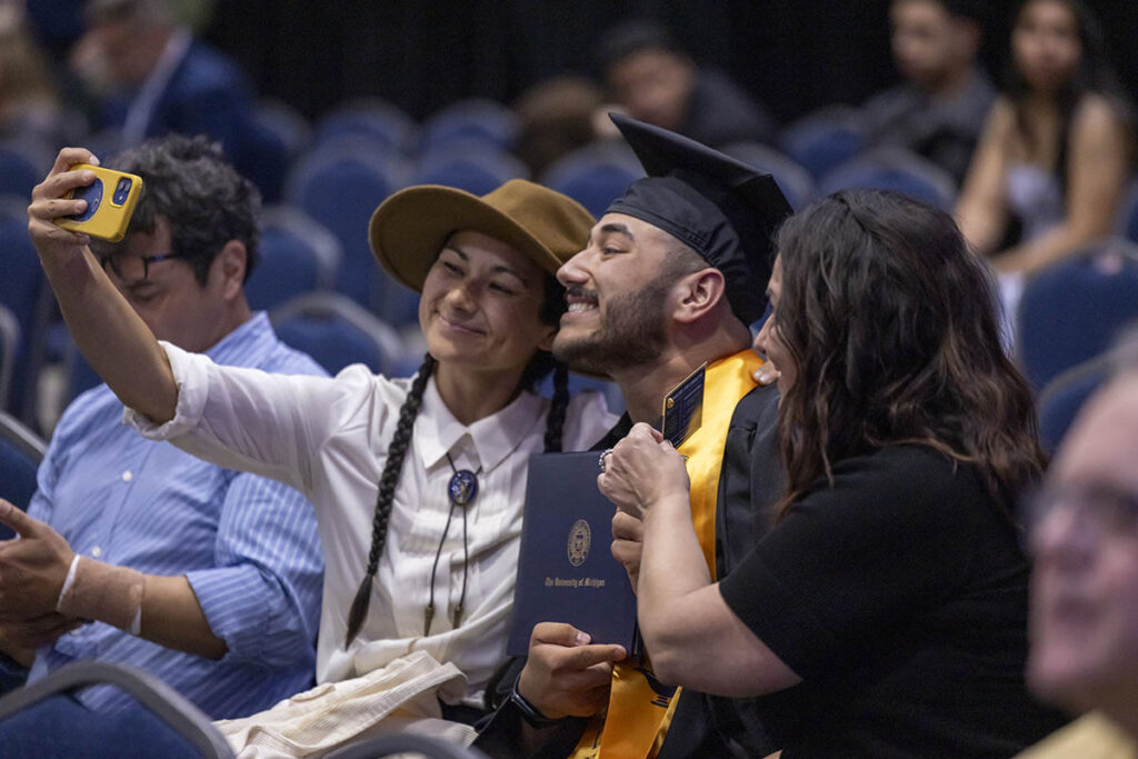 A male graduate sitting in the crowd with an older female and a younger female. The younger female is taking a selfie of the trio with a phone. The male graduate is wearing his graduation regalia and holding up his diploma.