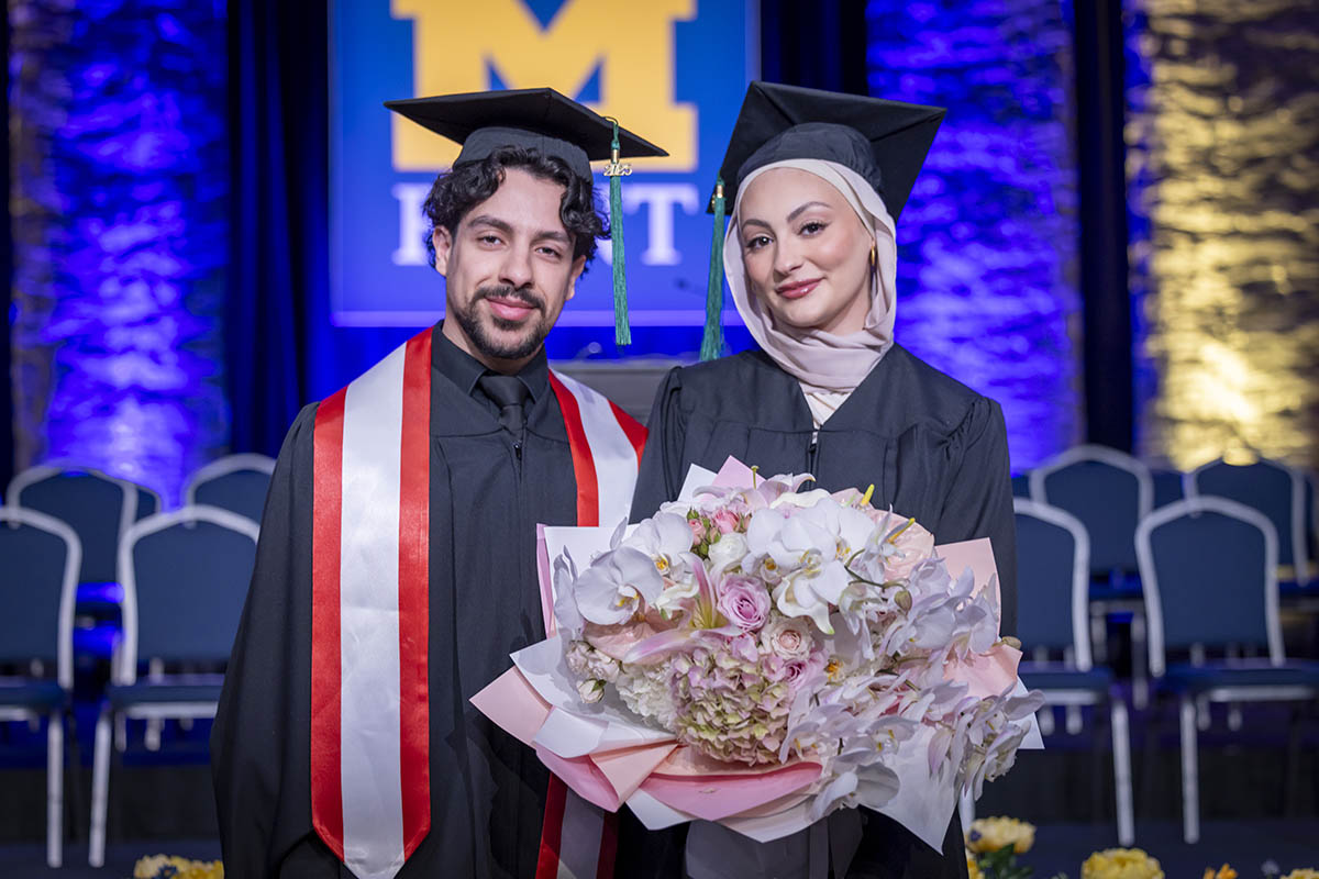 Two graduating students posing together. The female graduate has a bouqeut of flowers.