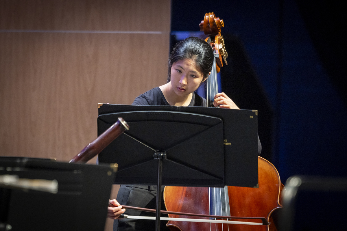 A young double bass player in black concert attire performs with a bow, reading intently from a black music stand, partially framed by nearby instruments.
