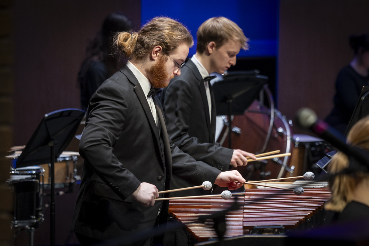 Two male percussionists in tuxedos concentrate as they perform on marimbas with soft mallets, with other percussion instruments and musicians in the background.