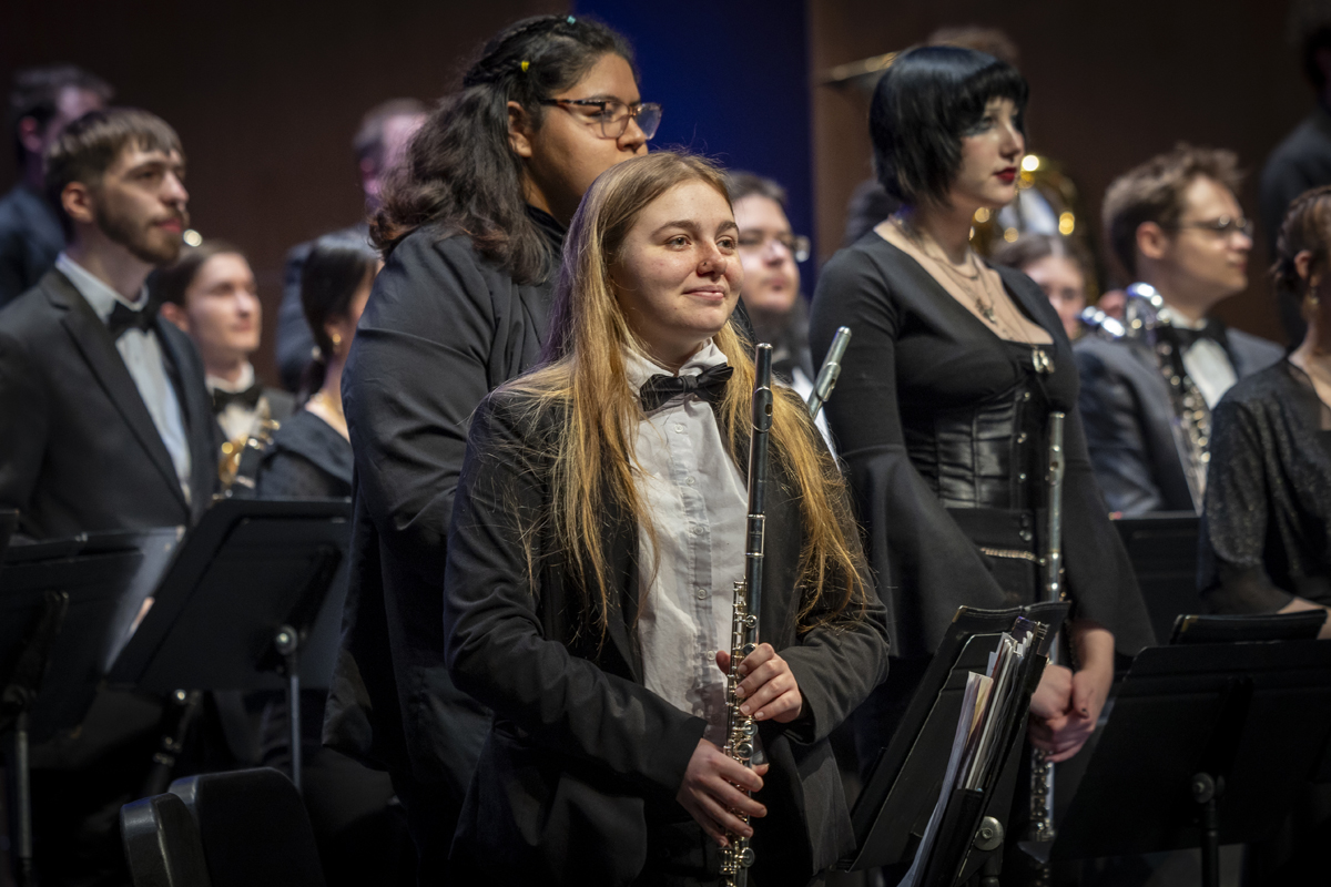 A group of musicians, including flutists in formal wear, stand with their instruments among rows of black music stands, appearing to acknowledge applause.