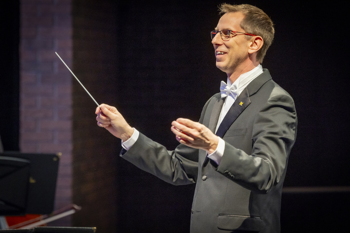 A male conductor in a tuxedo and white bow tie conducts with a baton, smiling and facing slightly to the right, with a dark background and part of a brick wall behind him.