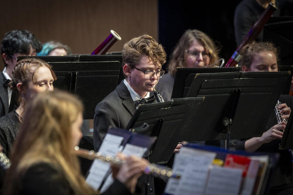A group of young woodwind players, including oboists and flutists, concentrate on their instruments during a live concert, surrounded by black music stands and dim lighting.