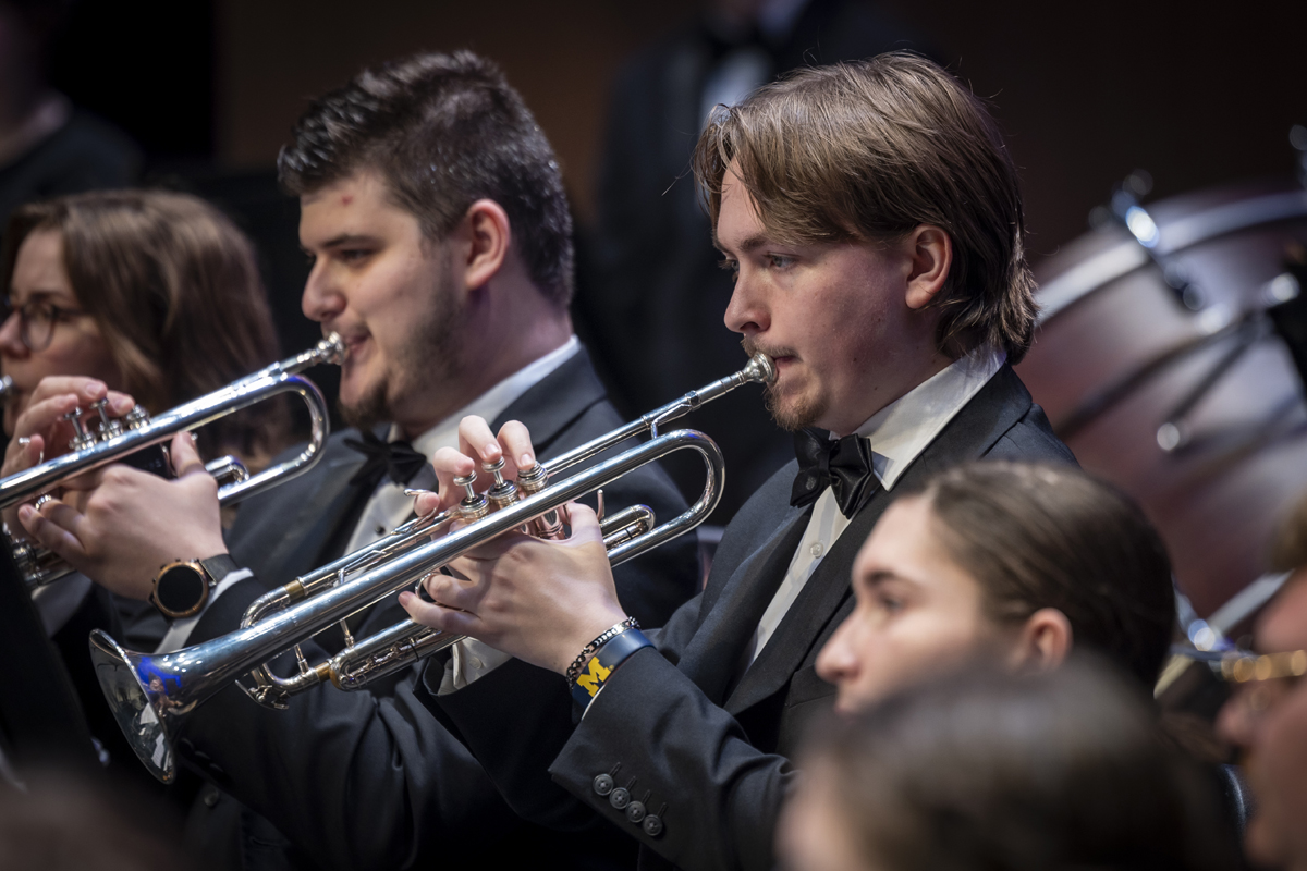 Two trumpet players in tuxedos perform in close coordination, focused on their instruments, with a bass drum visible in the background and other musicians partially in view.