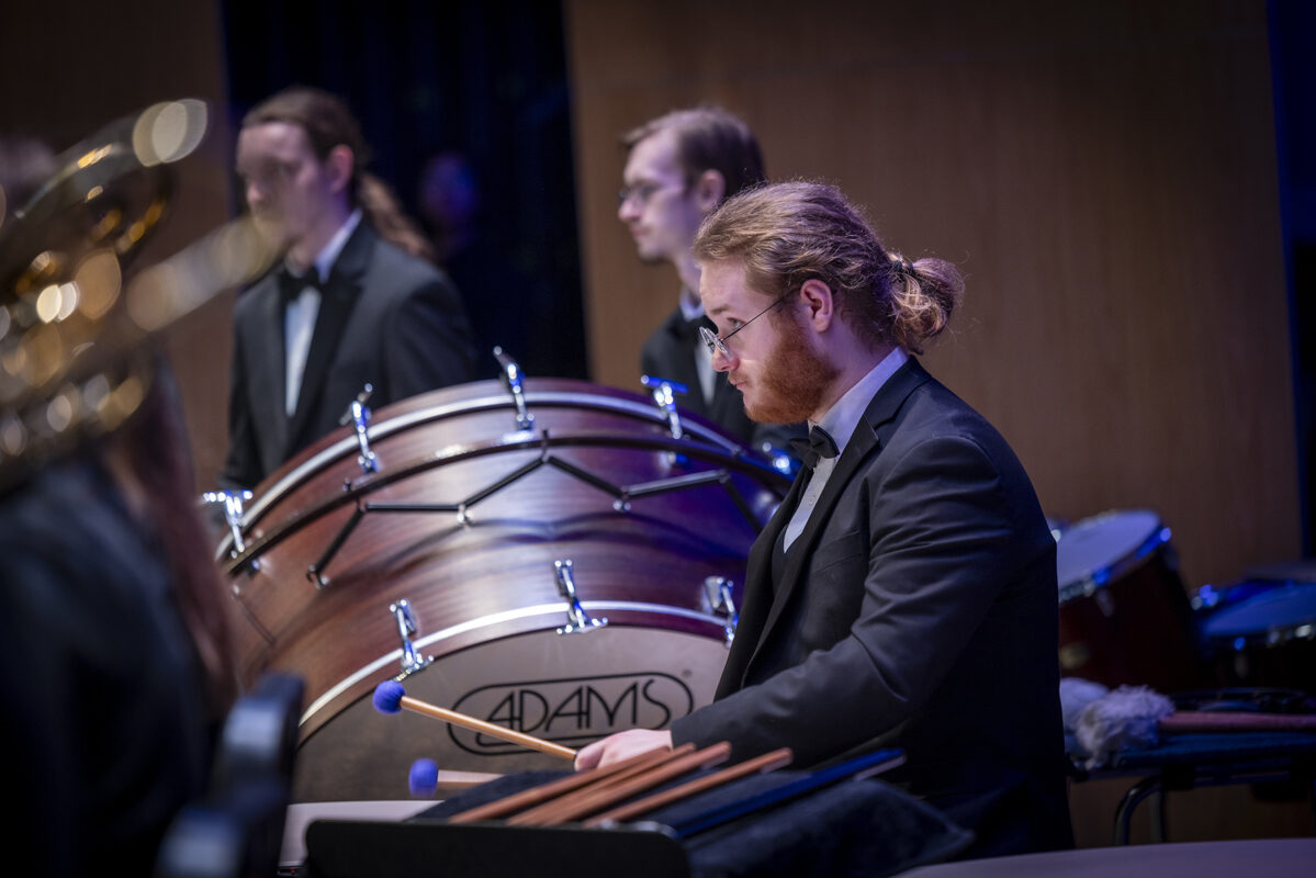 A percussionist in formal attire prepares to play with blue mallets at a concert, with a large bass drum behind him and other percussion instruments nearby.