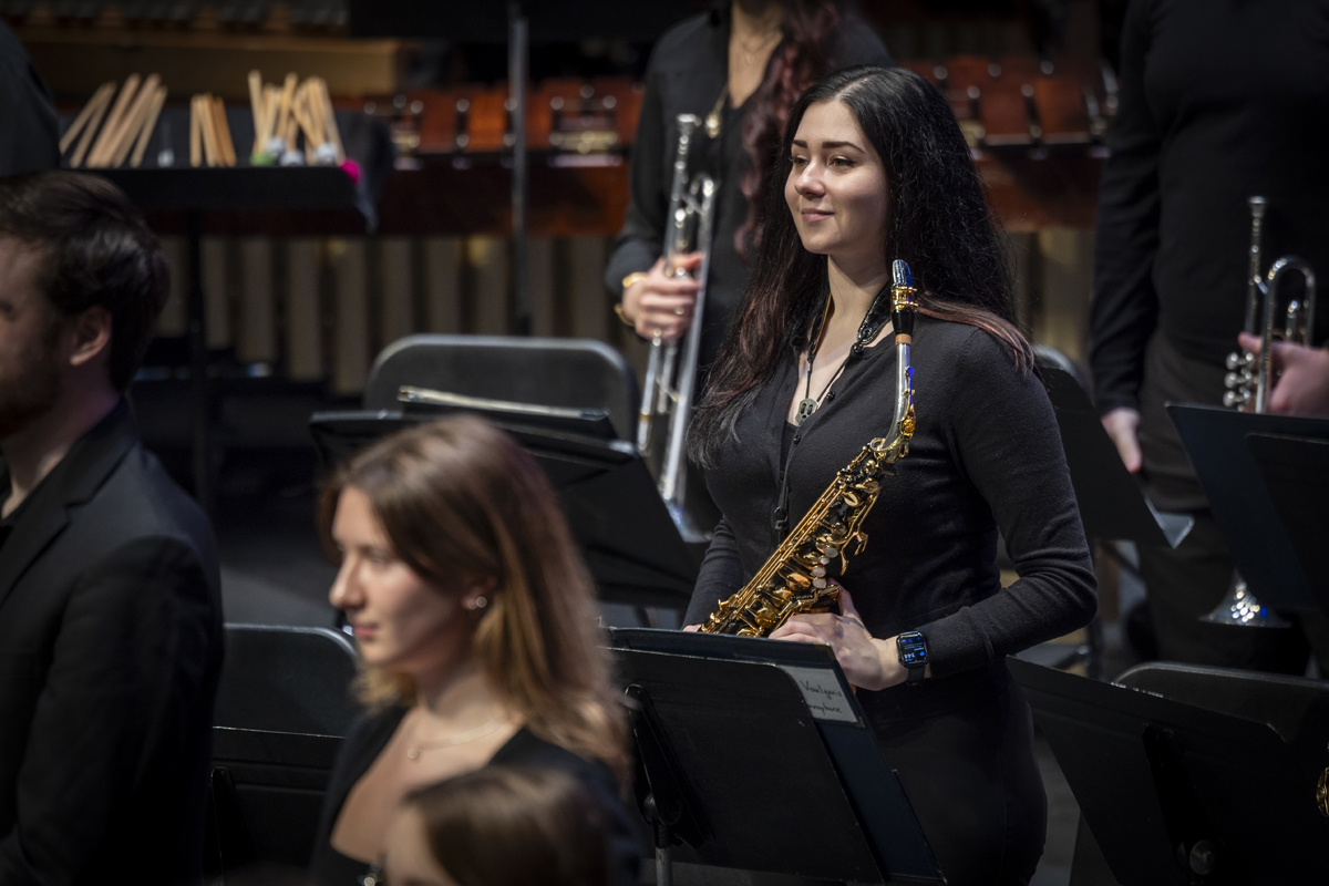 A young woman holding a saxophone stands confidently in a concert setting, smiling softly, with other musicians and percussion instruments behind her.