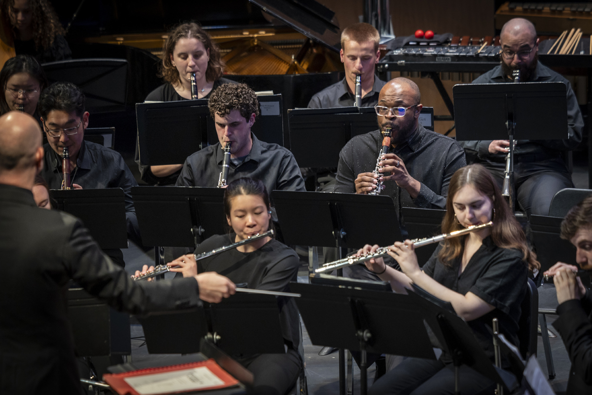 A group of woodwind players, including flutes, clarinets, and oboes, are seated in rows performing in a concert, while a bald conductor gestures in front of them.