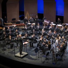 The University of Michigan’s Symphony Band composed of woodwind, brass, and percussion musicians dressed in black, performs on stage under a conductor in formal attire.