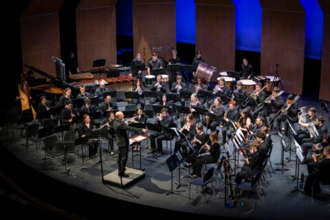 The University of Michigan’s Symphony Band composed of woodwind, brass, and percussion musicians dressed in black, performs on stage under a conductor in formal attire.