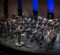 The University of Michigan’s Symphony Band composed of woodwind, brass, and percussion musicians dressed in black, performs on stage under a conductor in formal attire.