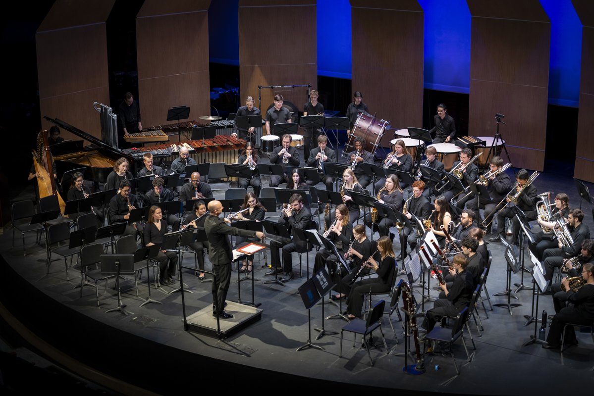 The University of Michigan’s Symphony Band composed of woodwind, brass, and percussion musicians dressed in black, performs on stage under a conductor in formal attire.
