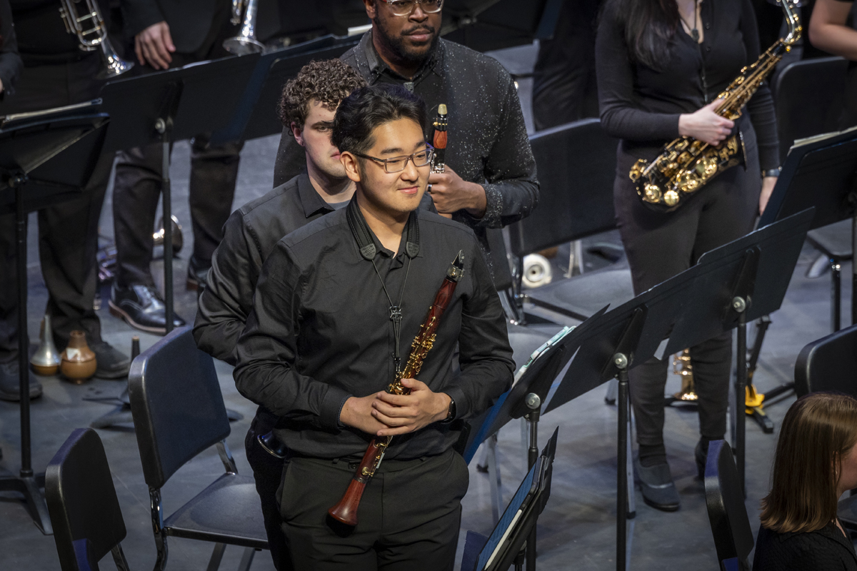 A group of symphonic band musicians stand among chairs and music stands, holding their instruments and smiling modestly, likely in response to audience applause.