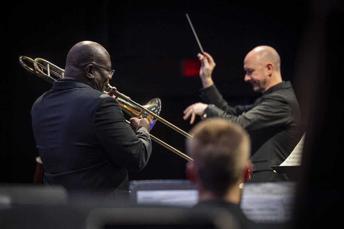 A trombone player stands in profile mid-performance, facing a conductor who gestures expressively with a baton, both dressed in black against a dark stage backdrop.