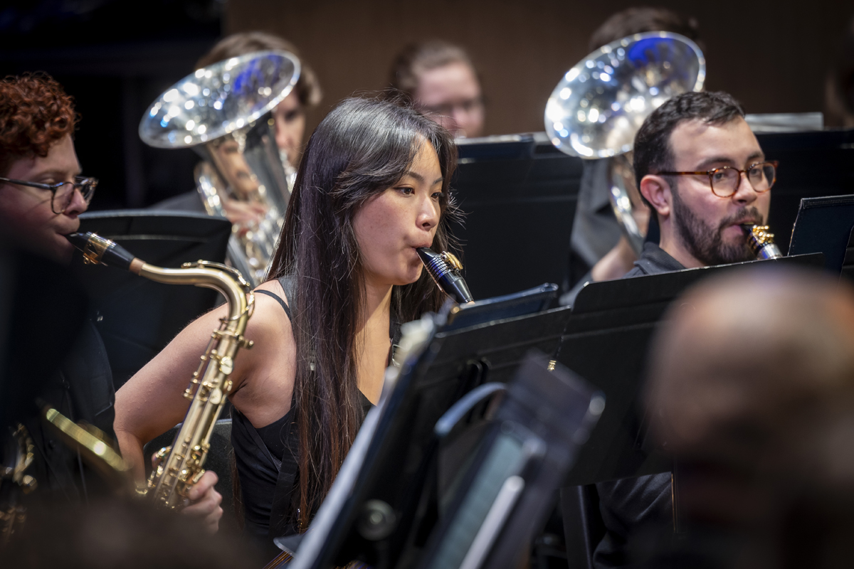 A close-up view of three concert musicians playing saxophones and clarinets, seated among black music stands, with brass instruments in the background.