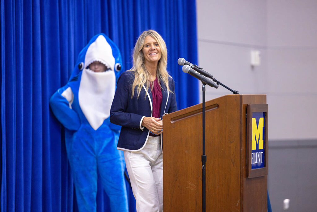 Paula Nas standing at a UM-Flint podium on stage with a shark mascot behind her.