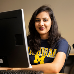 A woman with long, wavy dark hair and dark lipstick is seated at a desk smiling directly at the camera. She is wearing a navy blue t-shirt with the word "Michigan" in bold yellow letters above a large yellow "M," indicating the University of Michigan. In front of her is a large desktop monitor with a computer tower and keyboard partially visible. The setting appears to be an office or classroom with neutral walls and additional workstations in the background.