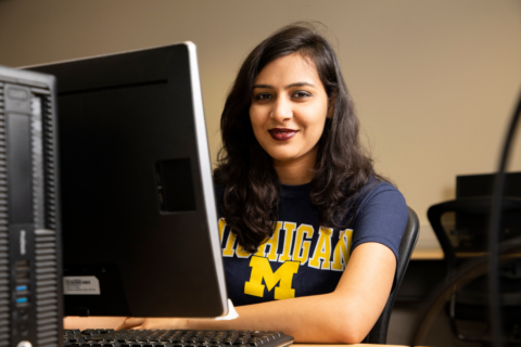 A woman with long, wavy dark hair and dark lipstick is seated at a desk smiling directly at the camera. She is wearing a navy blue t-shirt with the word "Michigan" in bold yellow letters above a large yellow "M," indicating the University of Michigan. In front of her is a large desktop monitor with a computer tower and keyboard partially visible. The setting appears to be an office or classroom with neutral walls and additional workstations in the background.