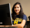 A woman with long, wavy dark hair and dark lipstick is seated at a desk smiling directly at the camera. She is wearing a navy blue t-shirt with the word "Michigan" in bold yellow letters above a large yellow "M," indicating the University of Michigan. In front of her is a large desktop monitor with a computer tower and keyboard partially visible. The setting appears to be an office or classroom with neutral walls and additional workstations in the background.