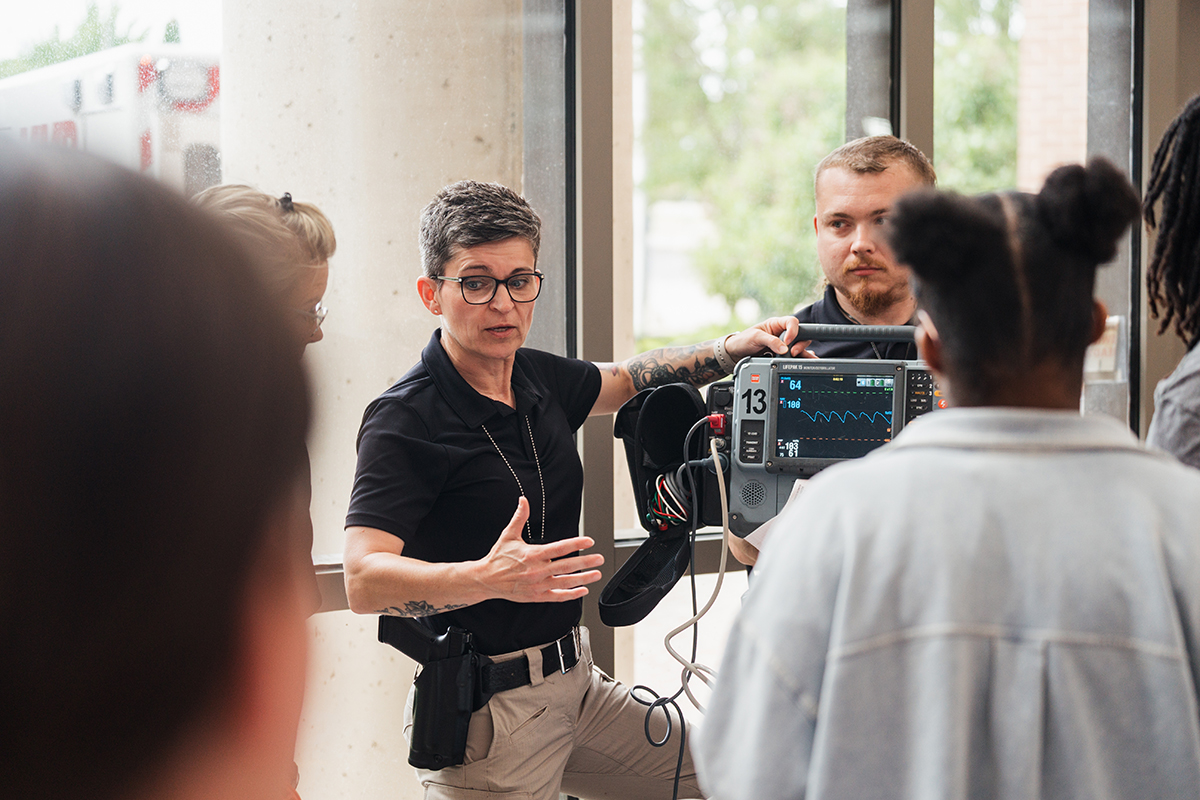 A female police officer standing next to a piece of electronic medical equipment talking to a crowd of K-12 camp attendees.