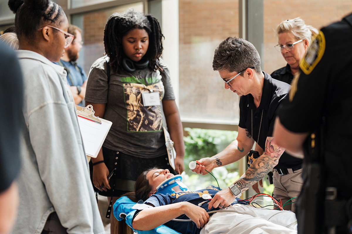 A female police officer attending to a mock patient on a stretcher while K-12 campers look on.