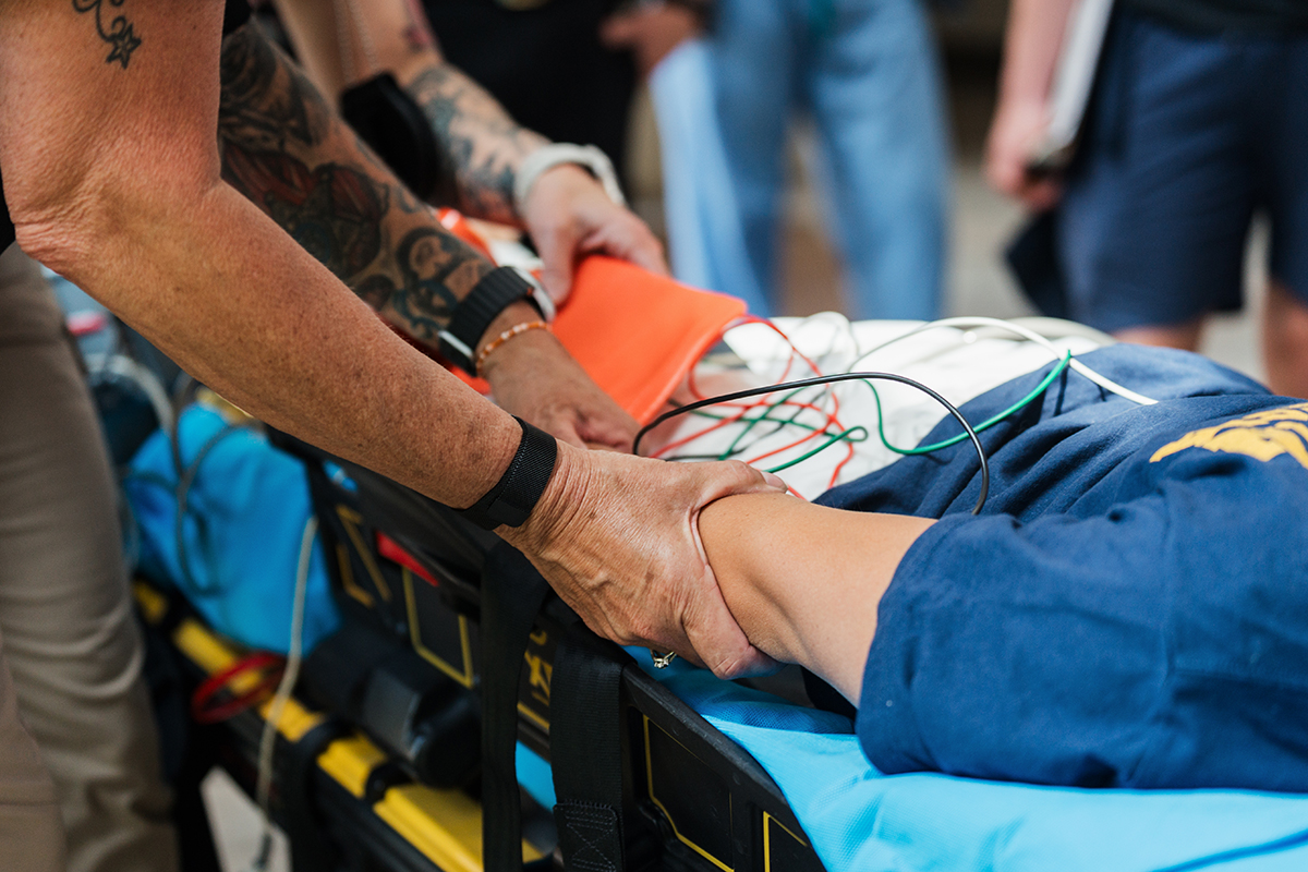 A close up of a mock patient's arm while the patient is on a stretcher and attached to some medical monitoring devices.