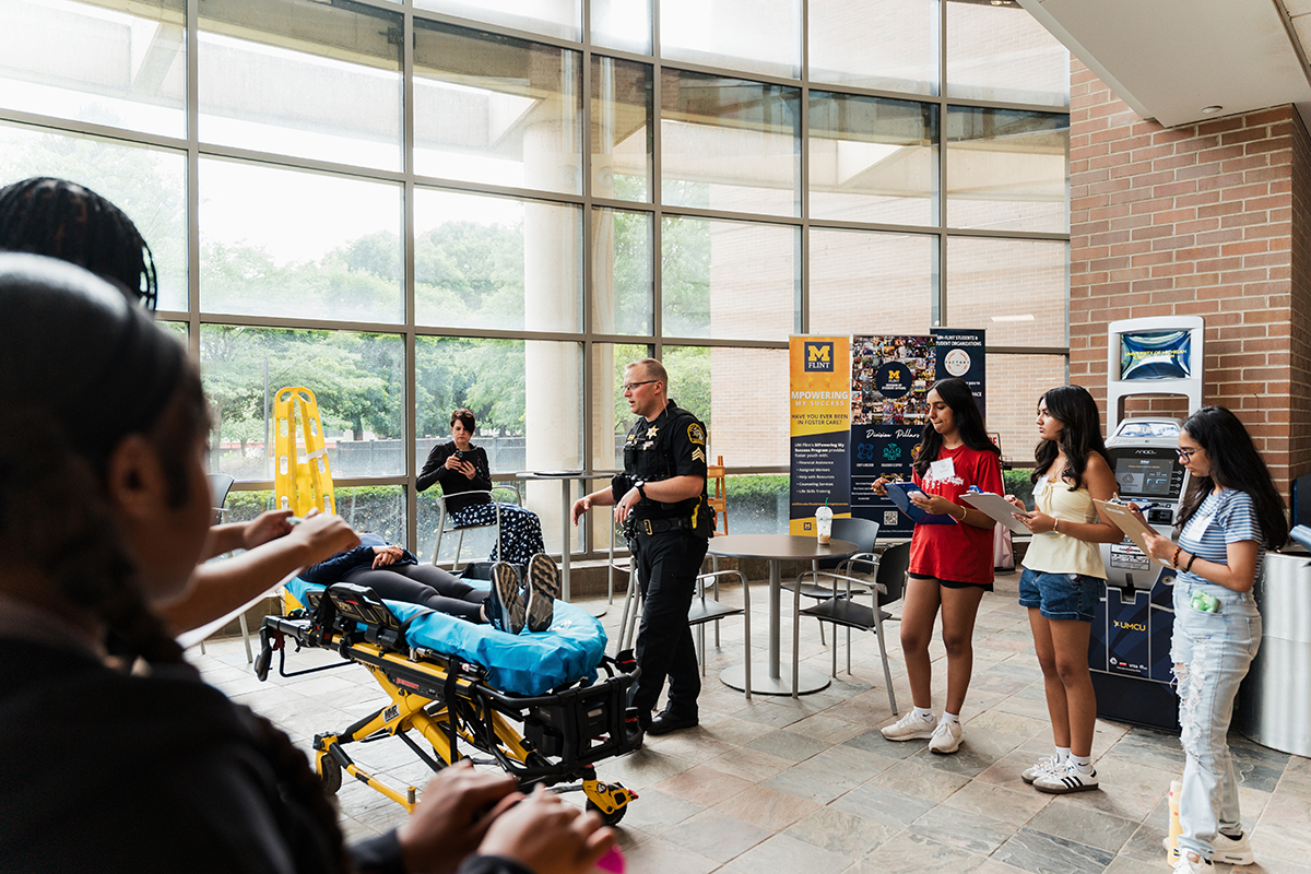 A male police officer speaking to the crowd in the William S. White Building while next to the mock patient on a stretcher.
