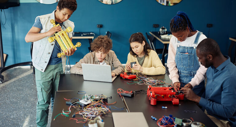 A diverse group of students building a robot with a teacher present.
