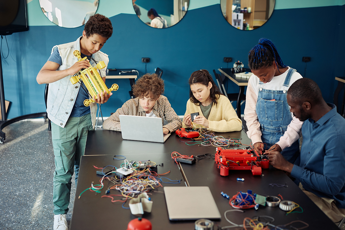 A diverse group of students building a robot with a teacher present.